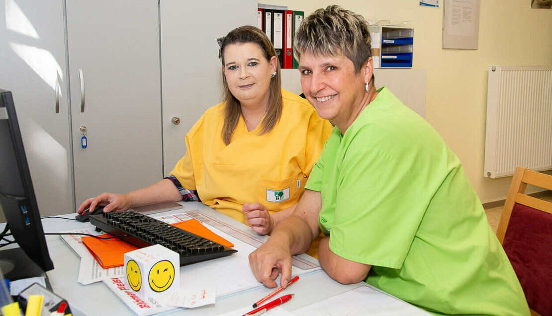 Zwei Damen in Arbeitskleidung der Pflege sitzen an einem Schreibtisch vor einem Computer und lächeln 