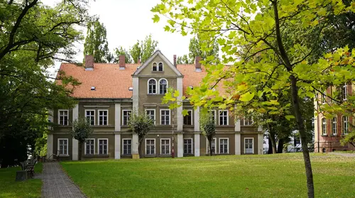 Ein dreistöckiges Gebäude mit Spitzdach umgeben von einer Parkanlage mit Rasenfläche und Bäumen