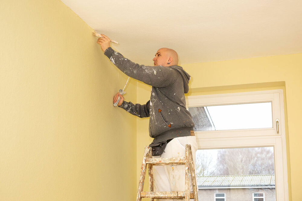 Ein Maler steht auf einer Leiter und streicht mit einem Pinsel die Kante zwischen Wand und Zimmerdecke