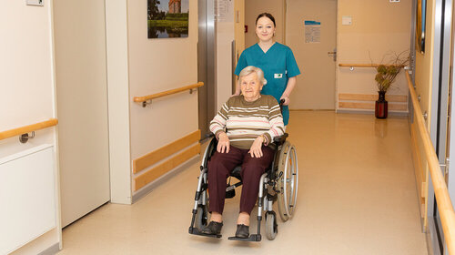 Eine junge Frau schiebt eine ältere Dame in einem Rollstuhl durch einen Flur