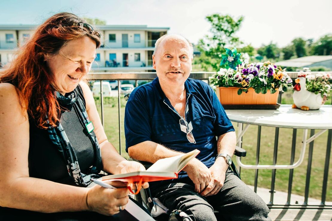 Ein Mann im Rollstuhl sitzt neben einer Frau auf seinem Balkon in der Sonne