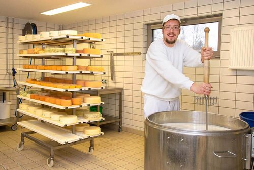 Ein Mann rührt mit einer Käseharve durch einen großen Behälter mit Milch. Im Hintergrund verschiedene frische Laibe auf einem großen Wagen.