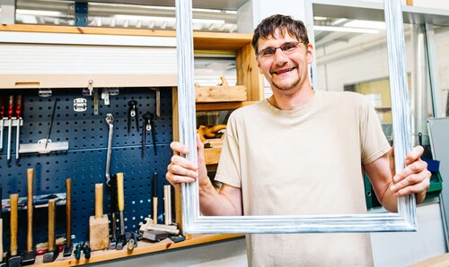 Ausschnitt aus unseren Lebenswelten Ein Mann in einer Tischlerei hält einen Fensterrahmen und blickt durch den Rahmen hindurch