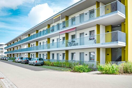 Außenansicht eines Wohngebäudes mit drei Stockwerken und Balkonen und davor parkenden Autos.