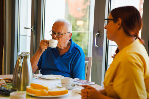 Ein älterer Herr nimmt eine Tasse Kaffee zu sich und eine Pflegekraft sitzt neben ihm am Tisch