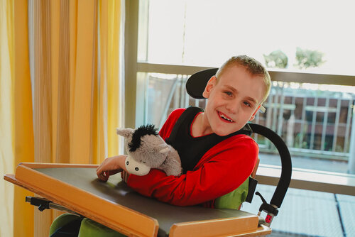 Ein Junge mit einem Kuscheltier im Arm sitzt in einem Rollstuhl und schaut in die Kamera