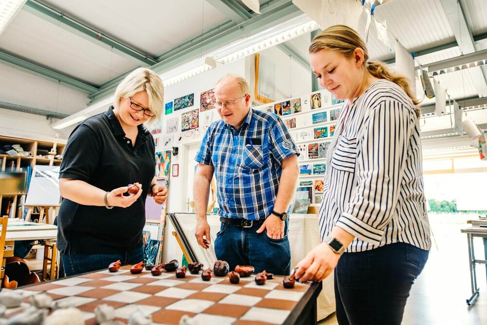 Zwei Frauen und ein Mann stehen um ein großes kariertes Brett, auf dem Figuren platziert sind