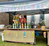 Ein bunt dekorierter Stand mit vielen Getränken und zwei Verkäufern, die den Daumen hochhalten