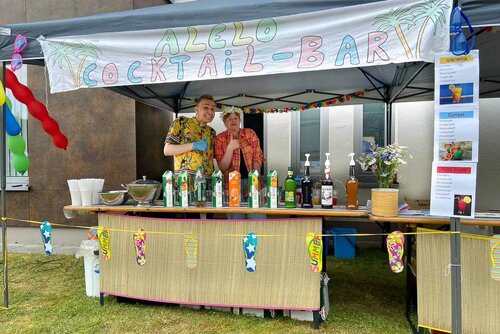 Ein bunt dekorierter Stand mit vielen Getränken und zwei Verkäufern, die den Daumen hochhalten