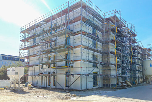 Im Bau sind Fenster eingesetzt und ein Gerüst steht um den Bau, man sieht nun eine glatte Hauswand