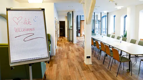 Ein Flipchart mit einem Willkommensgruß steht am Rand des Tagungsraumes mit einem langen Konferenztisch und vielen Stühlen.