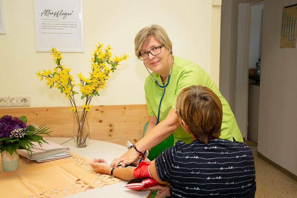 Eine Mitarbeiterin des Pflegedienstes Greifswald misst einer älteren Dame den Blutdruck und schaut dabei in die Kamera.