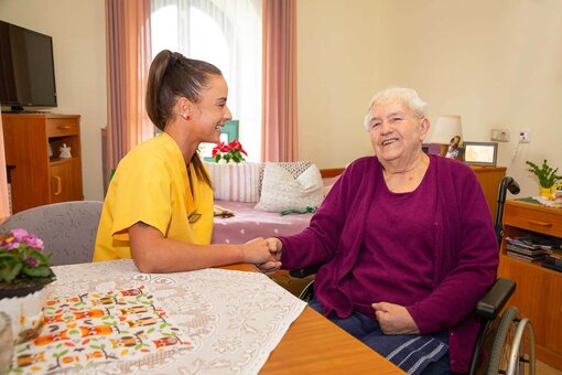 Eine junge Pflegefachkraft in Pflegekleidung hält einer älteren Dame im Rollstuhl die Hand und beide lachen