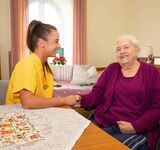 Eine junge Pflegefachkraft in Pflegekleidung hält einer älteren Dame im Rollstuhl die Hand und beide lachen