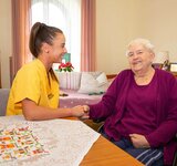 Eine junge Pflegefachkraft in Pflegekleidung hält einer älteren Dame im Rollstuhl die Hand und beide lachen