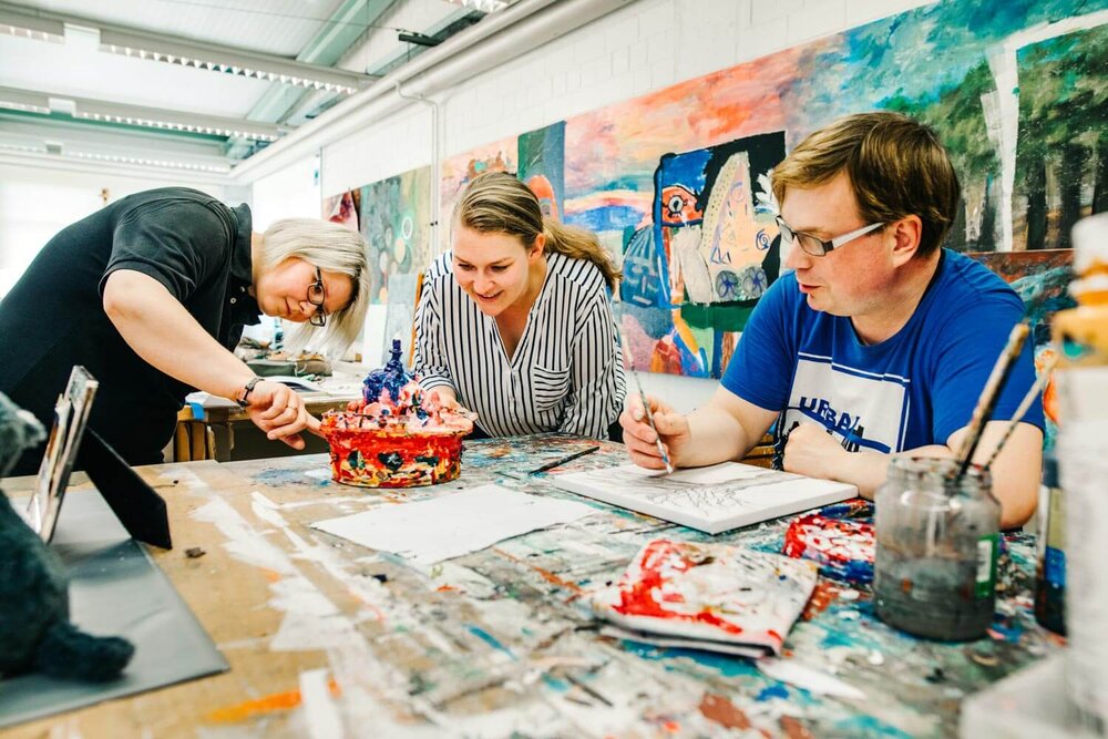 Zwei Frauen und ein Mann sitzen an einem Tisch und arbeiten an einem Kunstwerk mit Pinseln, Farben und Stiften