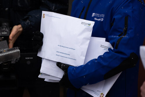 Ein Mann hält Umschläge in die Kamera, auf denen steht, dass es keine Werbung ist. In ihnen sind die Masken