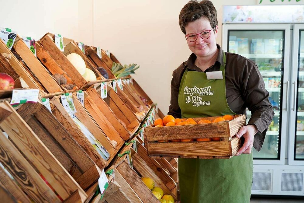 Eine Mitarbeiterin trägt einen Kisten mit Orangen zu einer Auslage mit frischem Obst und Gemüse im Pommerngrün