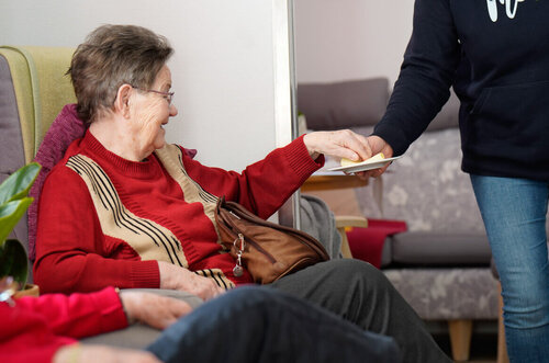 Eine ältere Frau sitzt in einem Sessel und bekommt von einer Pflegekraft einen Teller mit Keksen gereicht