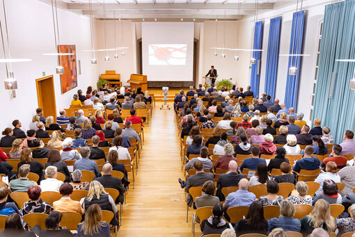 Ein großer Saal von oben mit vielen Stuhlreihen, in denen Menschen sitzen und am Ende ein Pult, wo Dr Bartels seine Rede hält