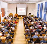 Ein großer Saal von oben mit vielen Stuhlreihen, in denen Menschen sitzen und am Ende ein Pult, wo Dr Bartels seine Rede hält
