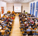 Ein großer Saal von oben mit vielen Stuhlreihen, in denen Menschen sitzen und am Ende ein Pult, wo Dr Bartels seine Rede hält