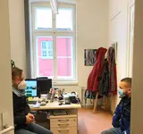 Ein Mann mit Maske sitzt gegenüber einer Frau mit Maske in einem Büro