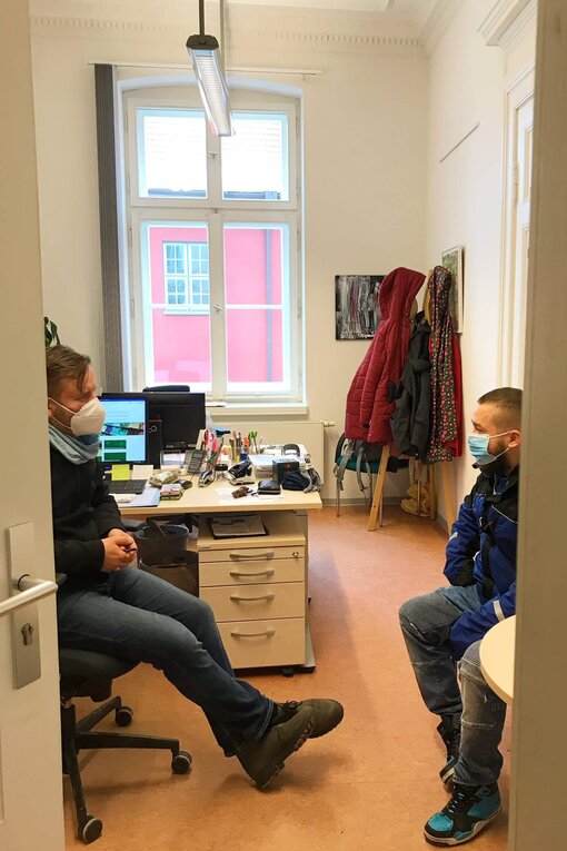 Ein Mann mit Maske sitzt gegenüber einer Frau mit Maske in einem Büro