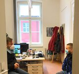Ein Mann mit Maske sitzt gegenüber einer Frau mit Maske in einem Büro