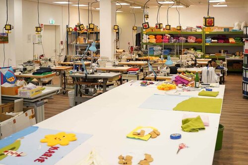 Die Näherei-Werkstatt mit vielen Werktischen und im Vordergrund einem großen Tisch mit Stoffen, Nähzeug und Mustern