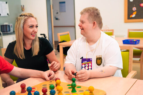 Eine Pflegekraft sitzt mit einem jungen Mann an einem Tisch und beide spielen Mensch ärgere dich nicht