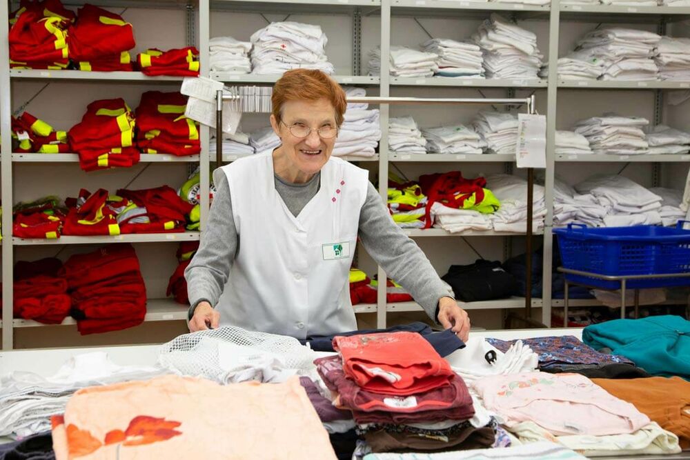 Eine ältere Dame steht lächelnd vor einem Regal mit zusammengelegter Wäsche und faltet weitere Wäschestücke