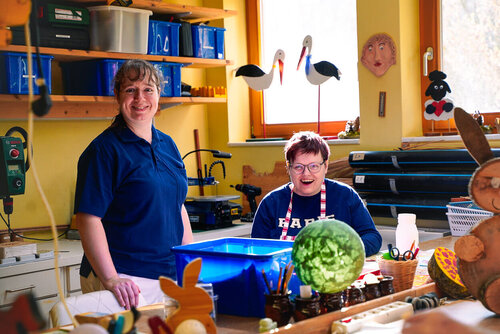 Eine Person mit einer Brille sitzt in Arbeitskleidung hinter einem Tisch und eine andere Person steht lächelnd neben ihr