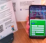 Ein Smartphone mit dem Check-In Formular des Hygiene-Rangers und ein erklärender Aufsteller