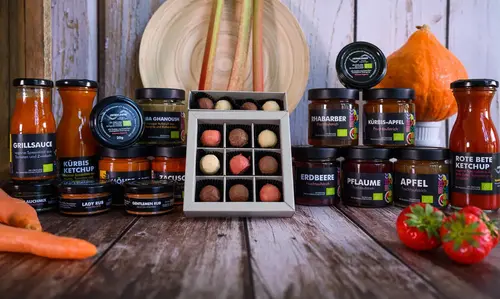 Gläser mit Aufstrichen, Pralinen und Fläschchen mit Saucen der Feinkostmanufaktur werden auf einer Holzoberfläche präsentiert.