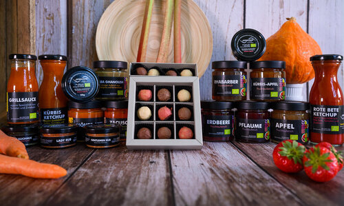Aus dem Sortiment der Feinkostmanufaktur Gläser mit Aufstrichen, Pralinen und Fläschchen mit Saucen der Feinkostmanufaktur werden auf einer Holzoberfläche präsentiert.