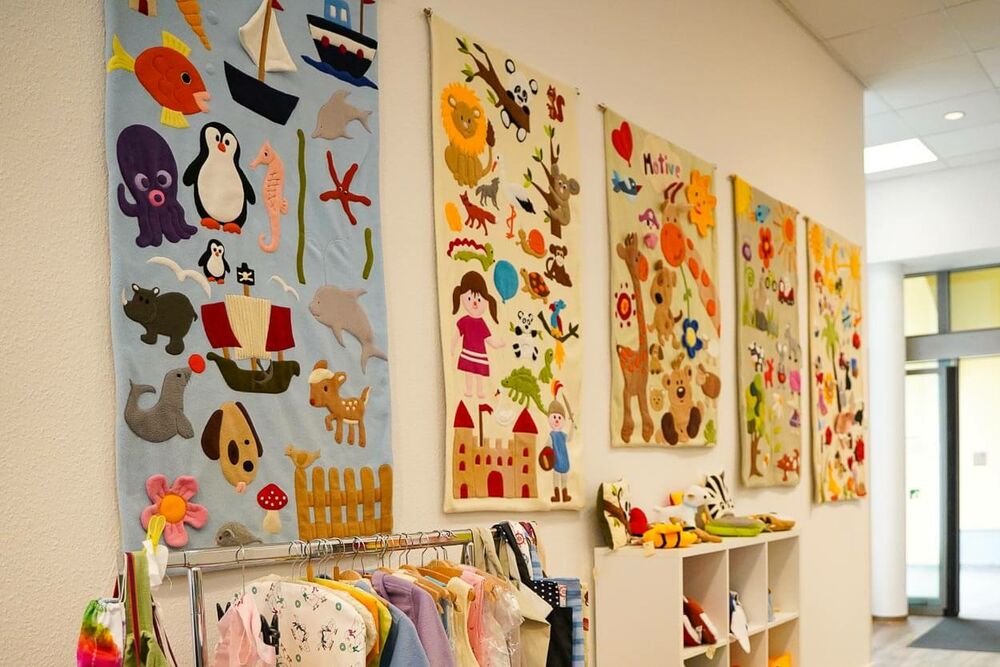 An einer Wand hängen mehrere bunte Babydecken aus Stoff mit Tiermotiven und mit anderen Motiven