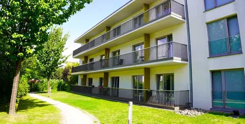Ein Gebäude mit drei Stockwerken und Balkonen, davor ein Fußweg und eine Parkanlage mit Rasen und Bäumen